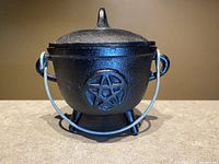 Closed view of cast iron cauldron with lid and pentacle symbol attached to a metal handle and standing on three short legs.