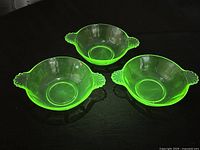 Three green uranium glass dessert bowls displayed upright on black surface, showing their translucent green color and scalloped handles.