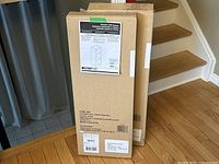 Two boxed units of ClosetMaid 3 Shelf Organizer standing upright on wooden floor near stairs.