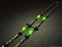 Close-up of the necklace showing four glowing neon green uranium glass beads set on a bronze-tone metal chain with smaller beads in between, glowing brightly under black light.