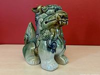 Front angled view of ceramic Foo Dog statue showing detailed face, mane, front paws and tail with blue and green glaze.