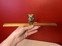 Hand-held vintage solid brass ruler with brass owl figurine on top, shown against a red background.