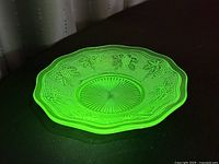 Green uranium glass bowl glowing under blacklight, showing grape and vine patterns and scalloped edge.