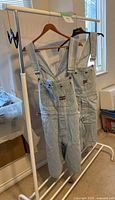 Two pairs of men's light blue denim Bugle Boy shortalls hanging on a white metal rack, showing the front and side views, one with a missing part on a buckle.