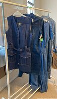 Four denim clothing items on hangers displayed on a rack: jumpsuits and dresses in different shades of blue denim.