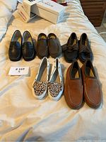 Five pairs of assorted Sperry loafers and boat shoes displayed on a bed, showing various styles including black and brown loafers, leopard print slip-ons, and boat shoes.