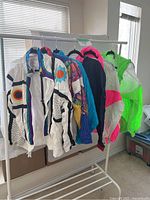Six vintage style track jackets hanging on a white clothing rack near a window, showing variety of colors and styles.
