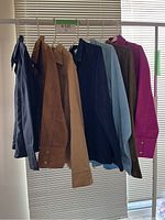 Seven vintage Western style shirts hanging on plastic hangers, showing a range of colors from dark navy, tan, light blue, olive, to magenta.