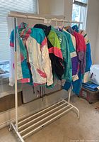 Seven vintage track suit jackets hung on a white garment rack, showing various bright colors of teal, pink, white, black, green, purple.