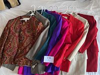 Nine vintage women's blazers in various solid colors and paisley pattern on hangers laid out.