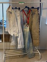 Full view of the four clothing items hanging on a white rack: a white denim jacket, white jeans, and light blue denim suspender jeans with red suspenders and coin trim.
