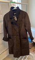 Front view of the long, dark brown sherpa coat hanging on a rack showing soft faux suede material, button-up front, two front pockets, and sherpa collar.