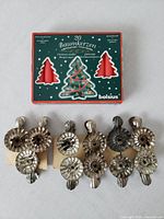 Set of 11 metal candle holders with adjustable clips arranged on table next to unopened box of 20 red Christmas tree candles