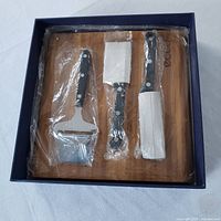 Cheese cutter set in original packaging on wooden board, showing four metal cutting tools with black wooden handles.
