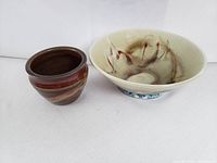Two hand-made pottery items side by side on white background: a larger bowl with abstract reddish and brown marks inside, and a smaller rounded pot with matte reddish glaze and beige stripes.