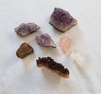 Overview photo showing all seven mineral and crystal specimens on a light cloth background, featuring three amethyst clusters, one citrine cluster, one quartz point, one rose quartz piece, and a dark fragment with gold flecks.