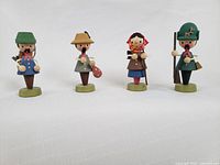 Four wooden figurines in a row on a white background showing full front view of each figurine spaced evenly