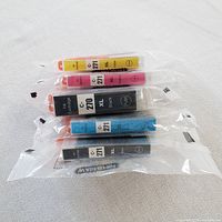 Five sealed Canon inkjet printer ink cartridges in yellow, magenta, black, cyan, and black colors, vacuum-sealed individually in plastic.