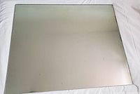 Photo shows large rectangular frameless wall mirror lying flat on a white surface, reflective surface with some slight marks and paint residue on edges visible.