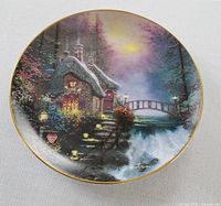 Front view of a collector plate titled Olde Porterfield Tea Room showing a stone cottage by a stream at dusk with lit windows and a bridge in the background, signed by Thomas Kinkade, gold rimmed.