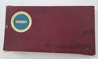 Maroon rectangular box with a circular blue and white 'Scrabble' label on top, corners showing wear and scuff marks.