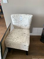 Accent chair with cream fabric and gray vintage script upholstery, showing front and partial side views on hardwood floor.