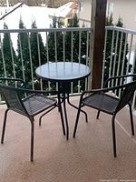 Outdoor bistro table with glass top and two black metal woven chairs on a balcony with a railing and distant mountain view.