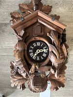 Front view showing the wooden cuckoo clock face with Roman numerals, surrounded by carved leaves, birds, and deer head with antlers.