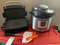 George Foreman grill with open grill plates alongside Instant Pot and orange food processor on red surface.