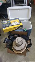 Open Coleman cooler with coiled hose inside, Martin cook stove box on side, hats and gloves around