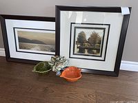 Two framed nature art prints and two leaf-shaped ceramic bowls in green and orange with a small fabric decorative piece on wood floor.