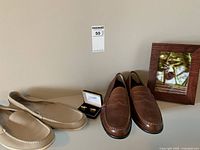 Pair of brown leather men's Rockport dress shoes placed on a shelf with other items including a photo frame and beige slip-ons.