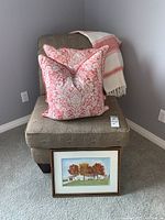 Armless chair in corner with two down pillows and blanket on top, framed picture leaning on chair base
