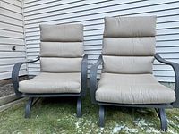 Two outdoor garden armchairs with dark grey metal frames and beige cushions placed outdoors on grass near a building.
