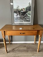 Full view of pine table with drawer and the framed artwork leaning on the wall behind it.