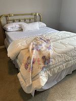 View of the entire bed with brass bedframe, mattress, pillows, comforter, and floral blanket draped on the side.