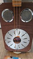 Close-up of the cigar box guitar body showing two metal sound holes and a round metal resonator plate with a central wooden circular hole and decorative cutouts.