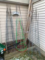 Several conical wire tomato cages/frames of varying heights in silver and a set in red and green against an outdoor siding wall.