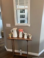 Photo of three white metal jugs and five colorful drinking mugs on a wooden table under a mirror.