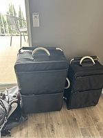 Photo showing 2 black fabric suitcases of different sizes with hard light-colored handles, placed indoors.