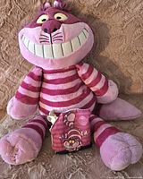 Full view of the plush Cheshire Cat Scentsy Buddy sitting on a blanket, with the matching scent pak in front.
