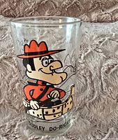Single clear glass tumbler with printed cartoon image of Dudley Do-Right in red Mountie uniform on one side, placed on textured fabric background.