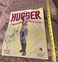 Large WWE Bayley signed poster lying flat on a blanket with a yellow background and "I'M A HUGGER" slogan, featuring Bayley in athletic outfit with boots and autograph in blue ink.
