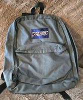 Front view of a medium gray JanSport backpack with logo patch and front zippered pocket