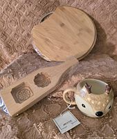 Photo shows a round wooden cutting board, a wooden mold with 2 floral carvings, and an animal-shaped ceramic mug with tag on a fabric surface.