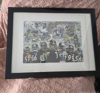 Framed print showing Pittsburgh Steelers players collage with black frame and white matting, lying on a pink textured surface