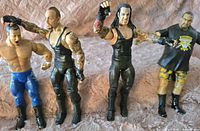 Four WWE wrestling figures displayed side by side on a pink fabric surface. From left to right: figure with blue pants and flexed muscles, Undertaker in black attire with tattoos, Stone Cold Steve Austin in black with tattoos, Road Dogg wearing a black t-shirt and camo shorts.