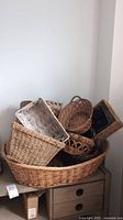 Photo showing multiple woven baskets stacked and arranged on a wooden dresser, including large oval baskets and smaller baskets of various shapes and colors.