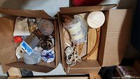 Two boxes with assorted items including coasters, drain strainers, decorative discs, bbs, glue sticks, metal legs, wooden parts, and miscellaneous hardware in a basement environment.