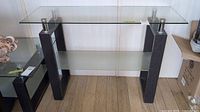 Full front view of the console table showing clear glass top, frosted glass shelf, wood legs and metal mounts.